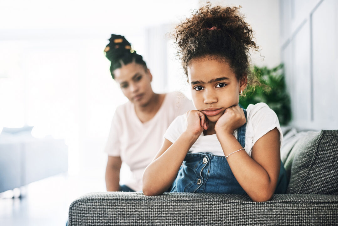 A young girl with a thoughtful, upset expression sits on a couch with her chin resting on her hands, while her mother looks on in the background with concern—illustrating a parenting moment where understanding a child’s temperament can improve communication and emotional support.