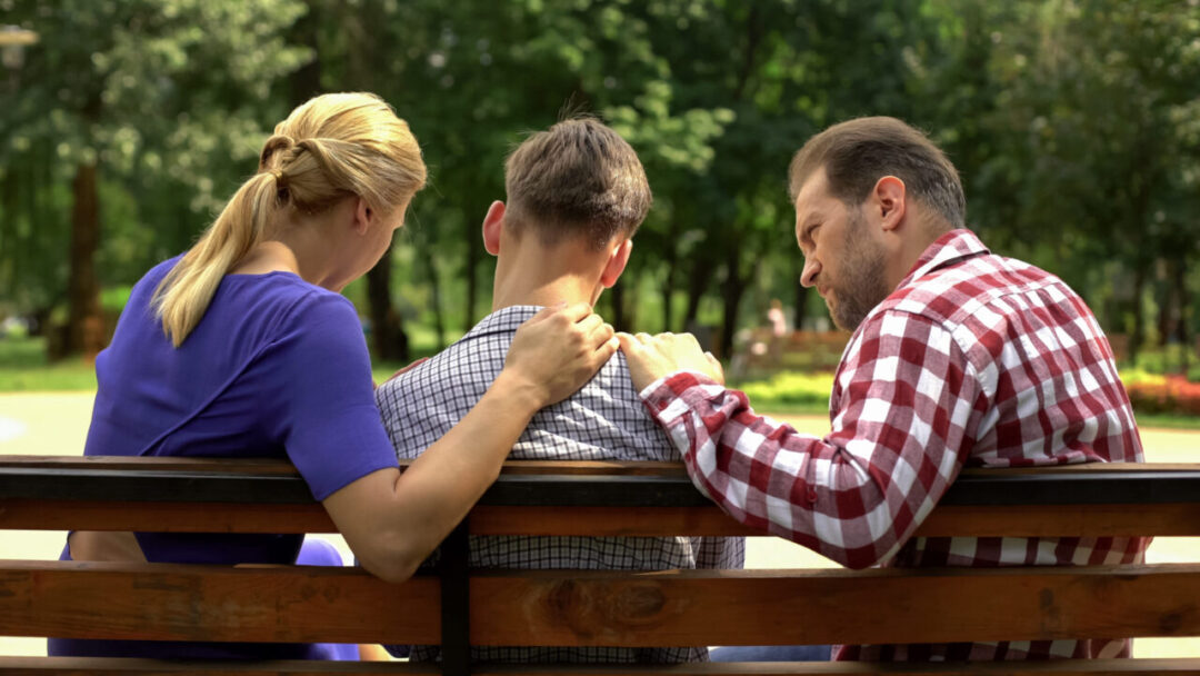 Parents sitting on a bench with their teenager between them, sharing a quiet, supportive moment together.