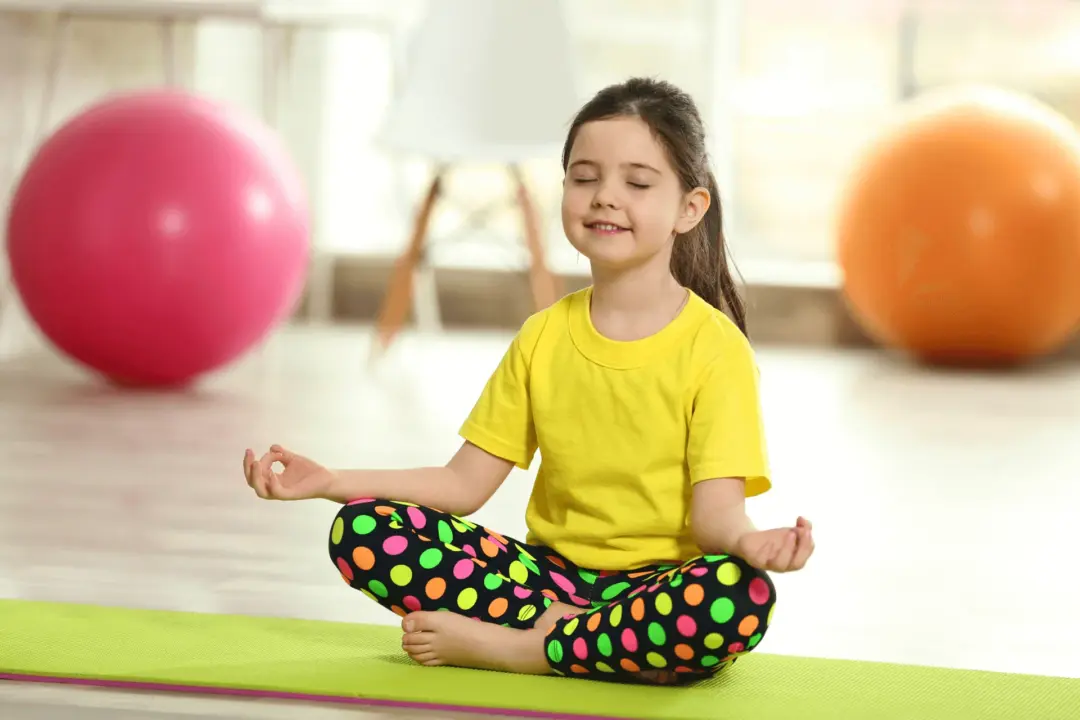 A young girl sits cross-legged on a yoga mat with eyes closed and a peaceful smile, practicing deep breathing or meditation in a bright room with colorful exercise balls in the background.