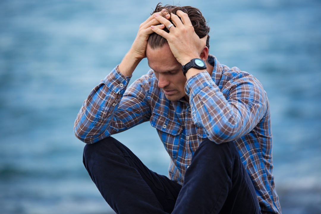 A man sitting by the water, holding his head in distress, wearing a plaid shirt and dark pants.
