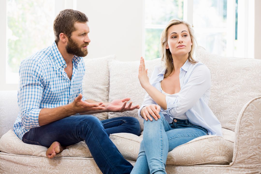 A couple sitting on a couch mid-argument; the man is expressing frustration with open hands while the woman raises her hand dismissively and looks away.