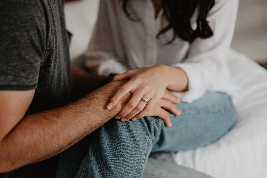 A close-up of a couple sitting closely together, with the woman gently holding the man's forearm in both hands. She is wearing a white blouse and blue jeans, and an engagement ring is visible on her finger, symbolizing emotional support and intimacy.