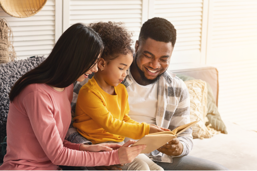 A smiling family sitting together on a couch, with parents and their young child enjoying a book, creating a warm and connected moment.