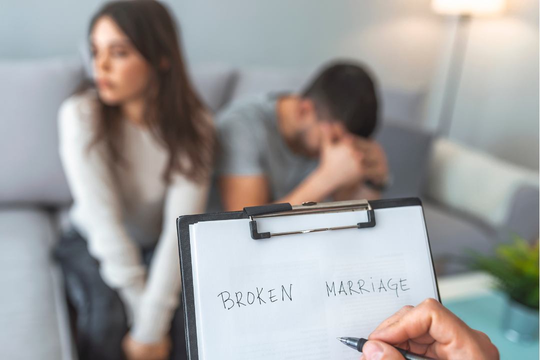 A distressed couple sits apart during a counseling session, with a therapist’s clipboard in the foreground reading “Broken Marriage,” symbolizing emotional disconnection and relationship strain.